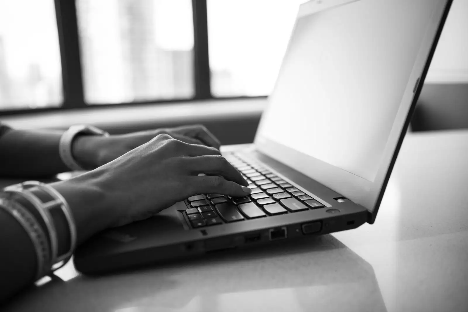 Gros plan en noir et blanc de mains tapant sur le clavier d'un ordinateur portable, près d'une fenêtre.