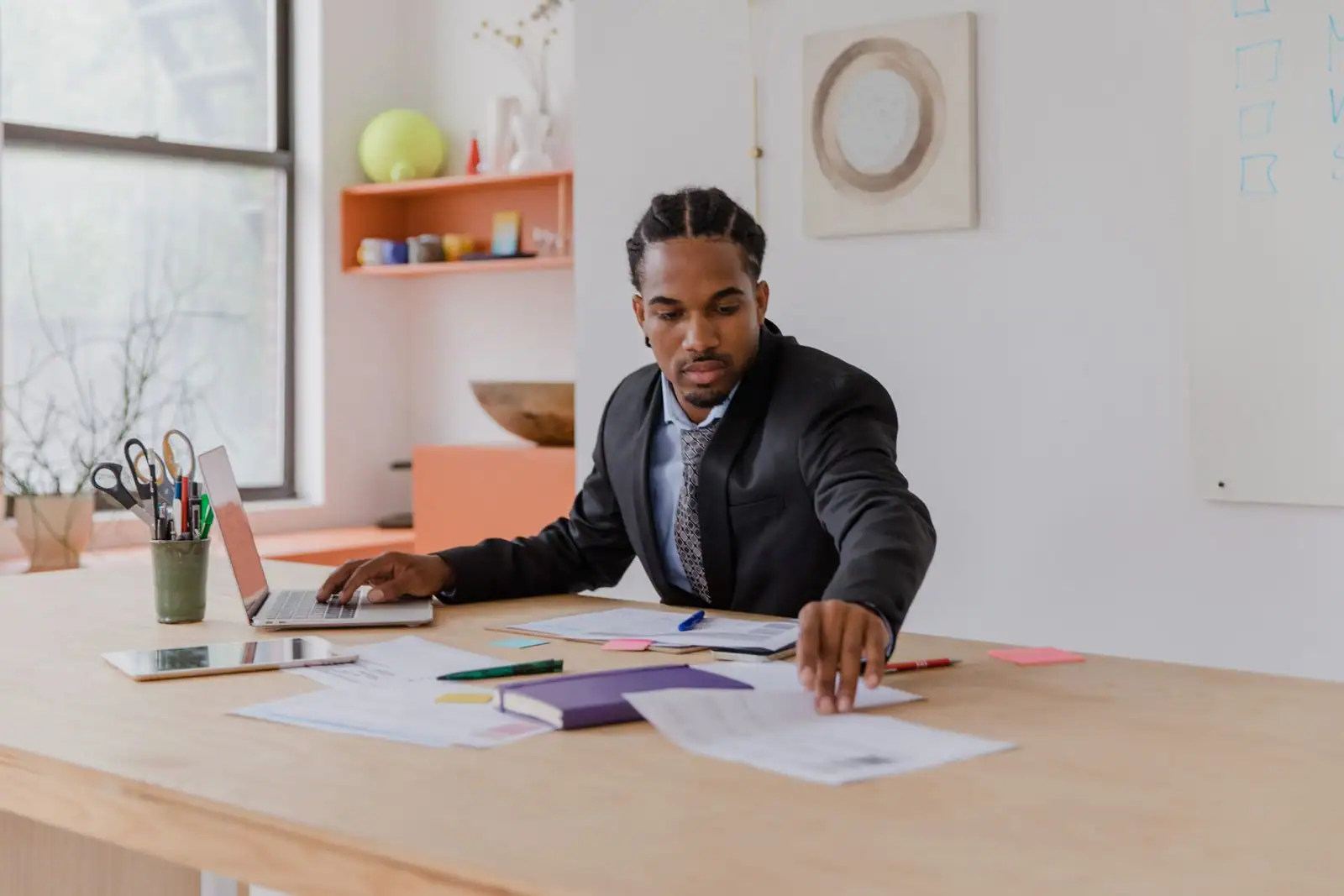 Un homme en costume travaille sur un ordinateur portable, entouré de documents posés sur un bureau, dans un intérieur lumineux.