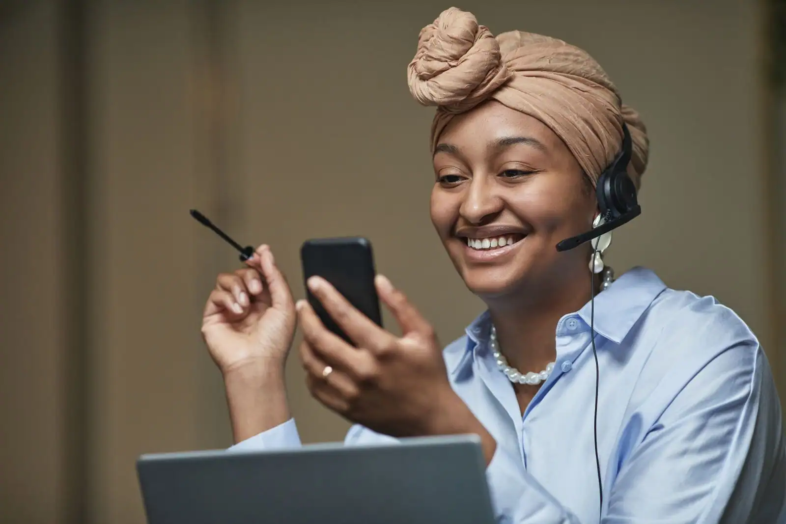 Femme souriante portant un casque-micro, smartphone en main, devant un ordinateur portable.