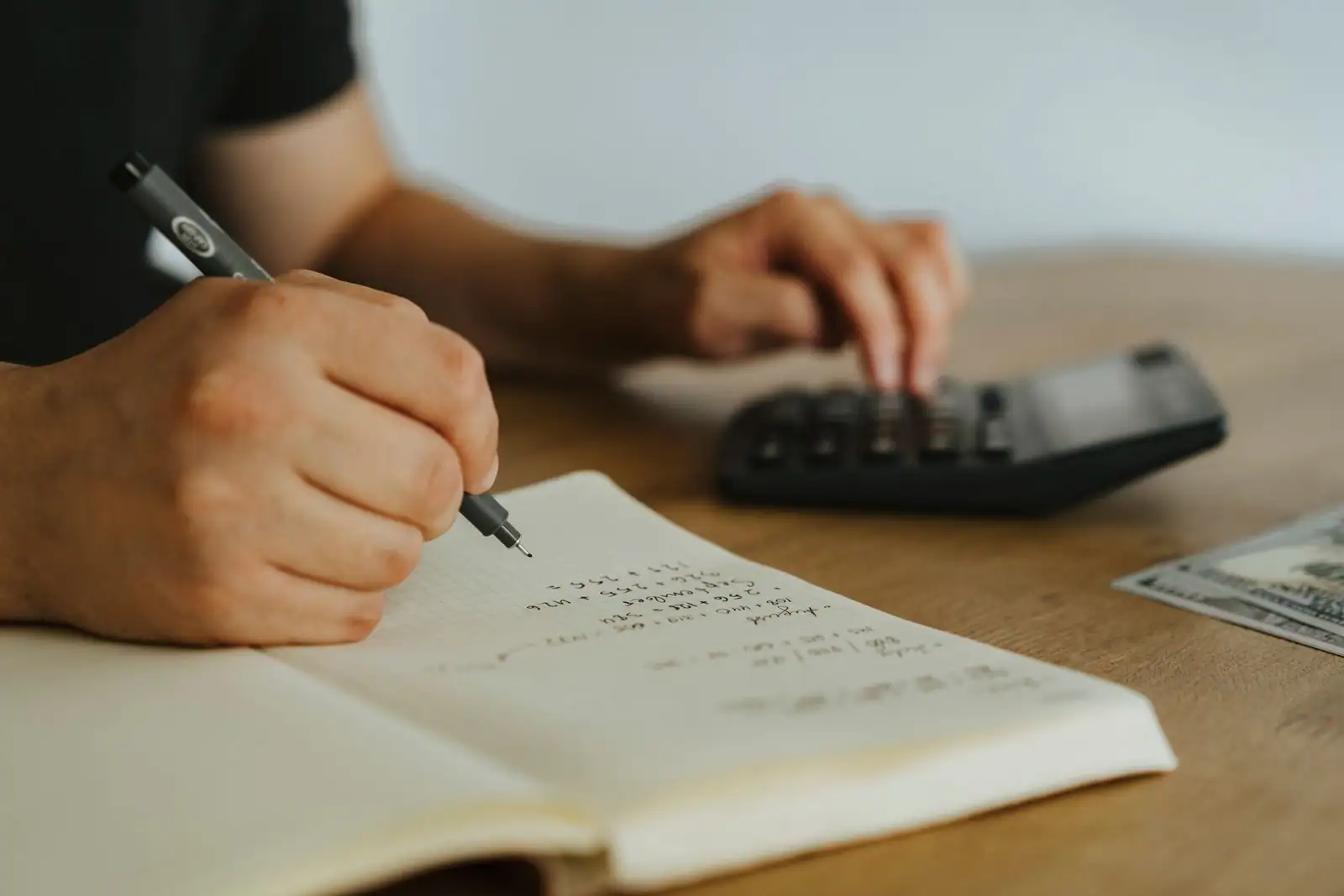 Personne calculant des prévisions financières à la main dans un carnet, avec une calculatrice, sur un bureau en bois