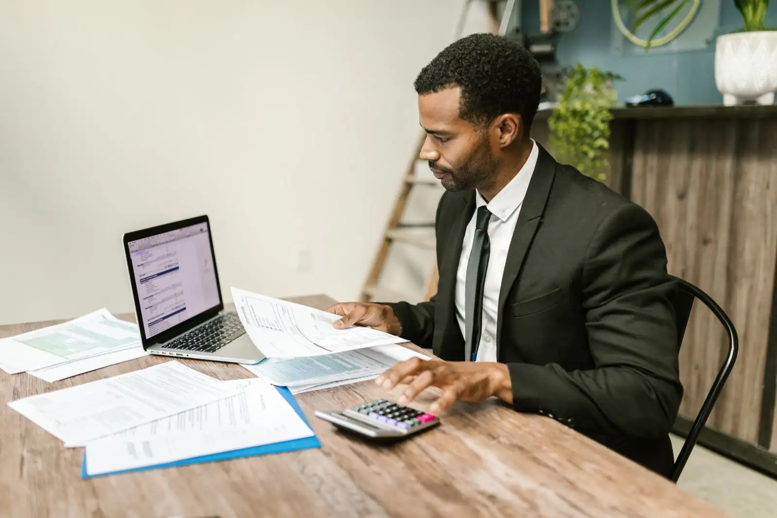 Dirigeant en costume examinant des documents comptables avec une calculatrice, un ordinateur portable posé sur son bureau.