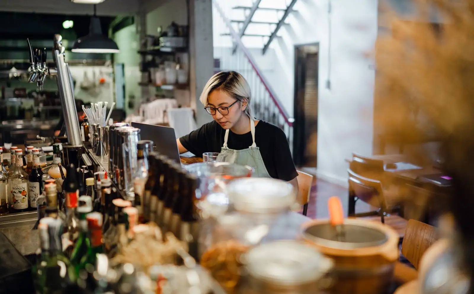 Une personne en tablier travaille sur un ordinateur portable derrière le comptoir de son bar.