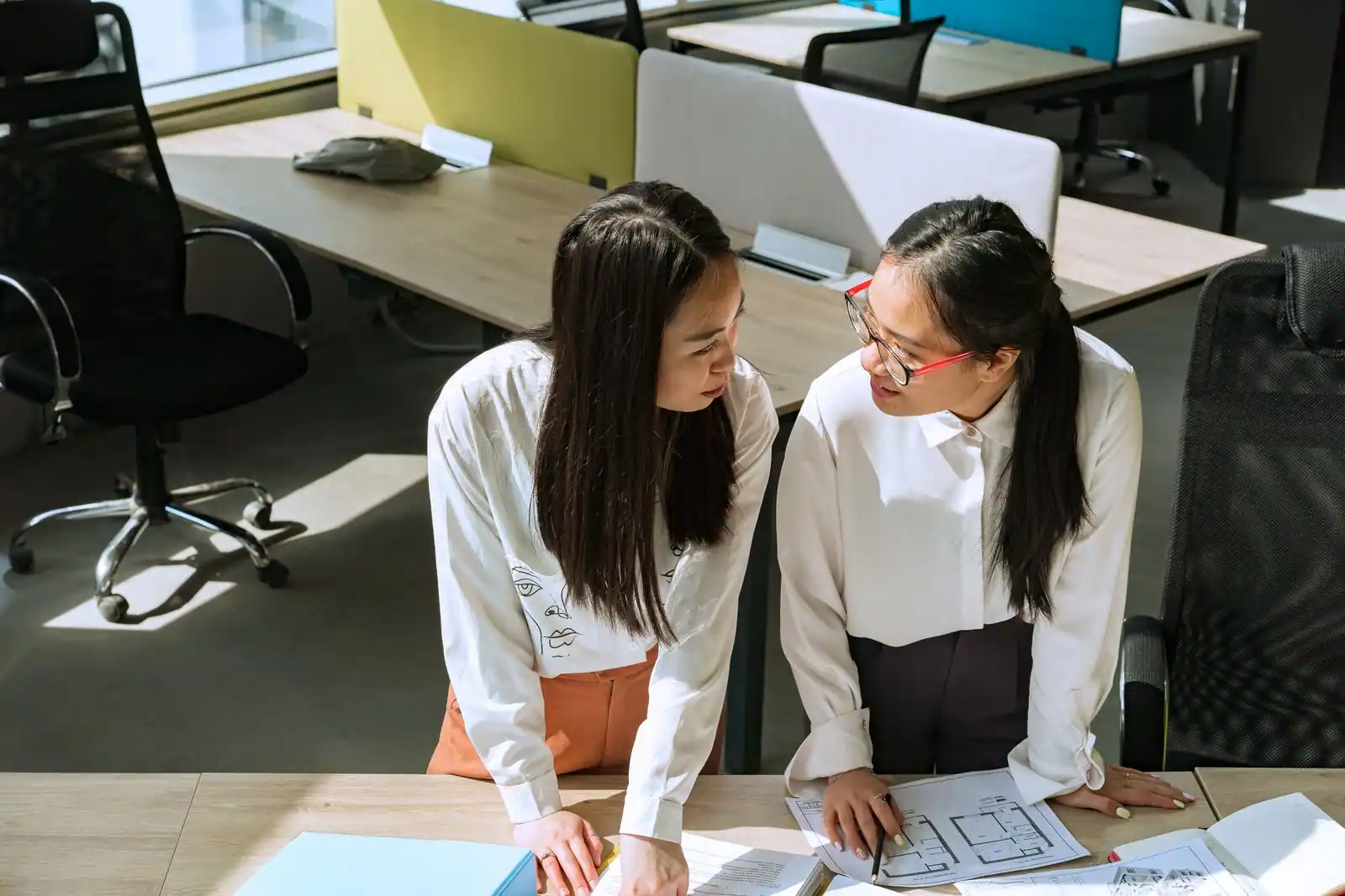 Deux collègues de même niveau discutent face à face, appuyées sur un bureau en open space
