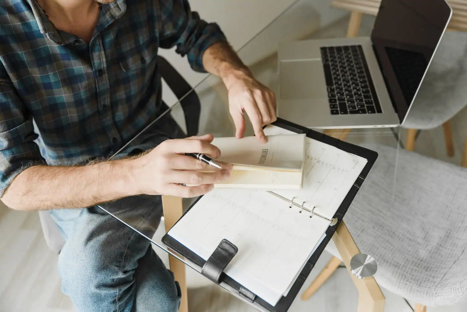 Un homme annote son agenda, un ordinateur portable ouvert à côté, sur un bureau en verre clair