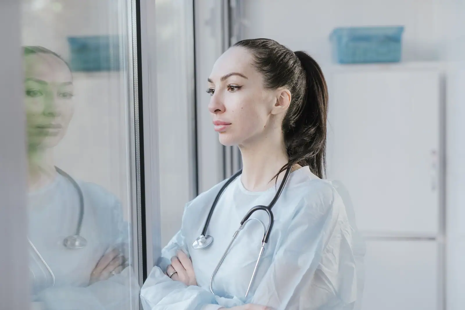 Soignante en tenue de protection, stéthoscope au cou, songeuse devant une fenêtre d'hôpital