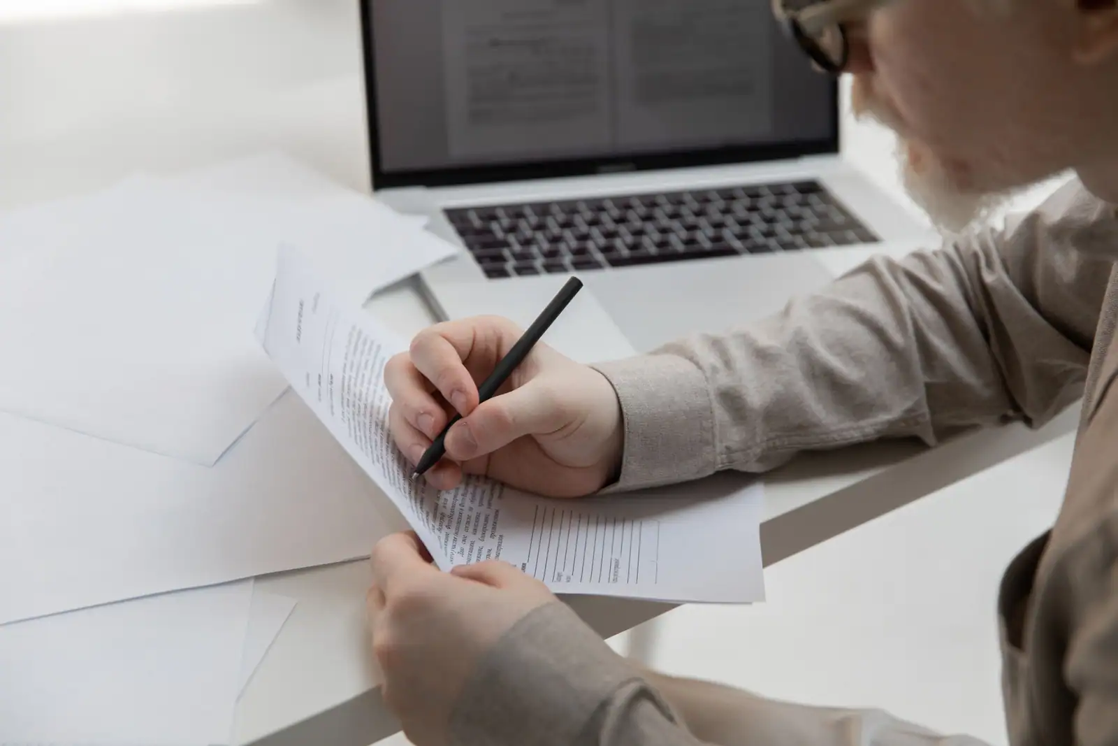 Mains annotant au stylo un document imprimé, devant un ordinateur portable ouvert sur un texte